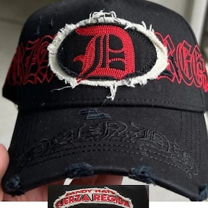 May include: A black baseball cap featuring a distressed, frayed-edge patch with a red letter D on the front. The cap displays red gothic-style text reading FUERZA REGIDA across the crown and brim, with additional frayed detailing on the visor edge.