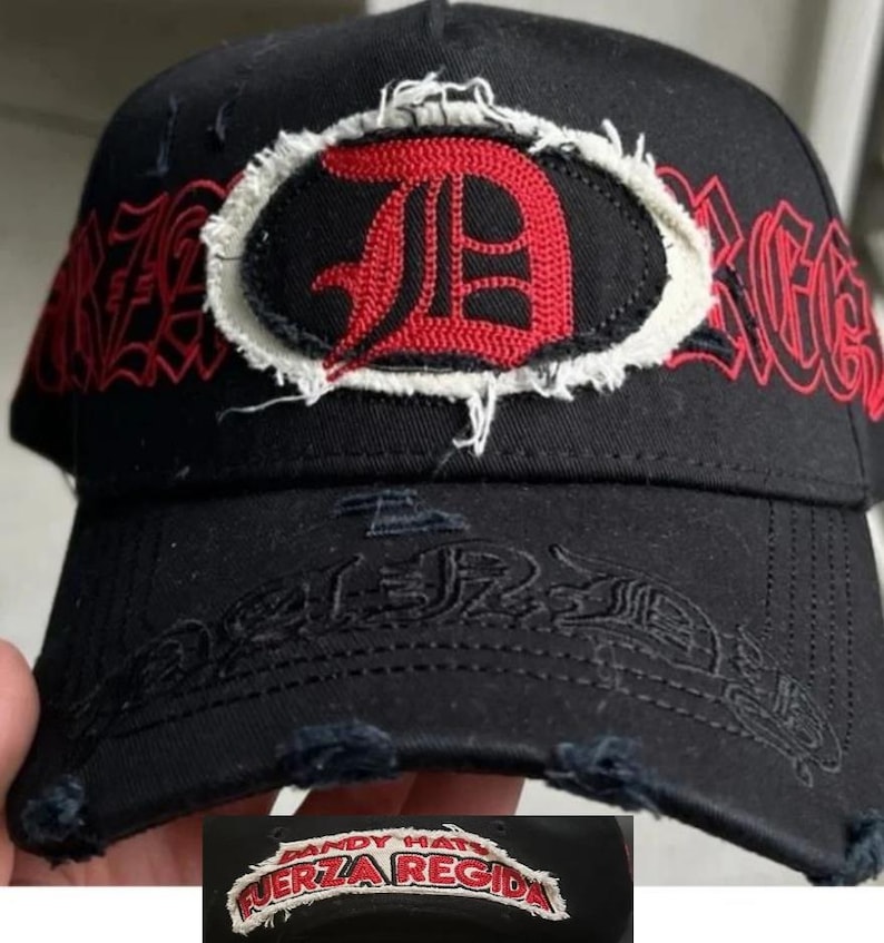May include: A black baseball cap featuring a distressed, frayed-edge patch with a red letter D on the front. The cap displays red gothic-style text reading FUERZA REGIDA across the crown and brim, with additional frayed detailing on the visor edge.