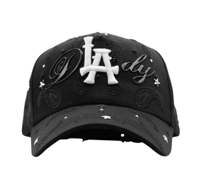 May include: Black baseball cap with white "LA" lettering and "dy" script. The cap features star-shaped embellishments and paisley patterns. A stylish accessory for casual wear.