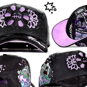 May include: A black baseball cap featuring an embroidered purple and black flower with a stitched face on the front. The cap has a textured black surface, a glossy brim, and a purple underside. The interior lining displays purple and black graphics.