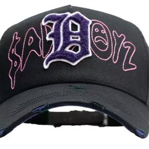 May include: Black trucker cap featuring the word "SABOIZ" embroidered in pink and purple. The letter "B" is in a purple patch with a white outline. The cap has a distressed brim and mesh back.