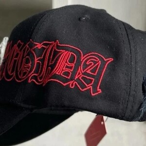 May include: A black baseball cap featuring red embroidered text in a gothic-style font across the side panel. The cap has a structured crown, a curved brim, and a small frayed fabric detail on the side, with a red tag hanging from the bottom edge.