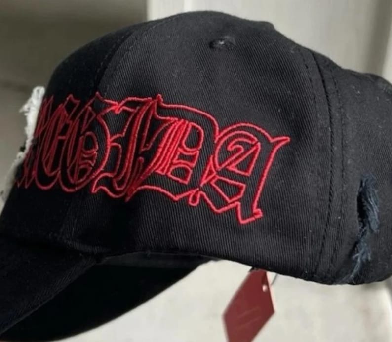 May include: A black baseball cap featuring red embroidered text in a gothic-style font across the side panel. The cap has a structured crown, a curved brim, and a small frayed fabric detail on the side, with a red tag hanging from the bottom edge.