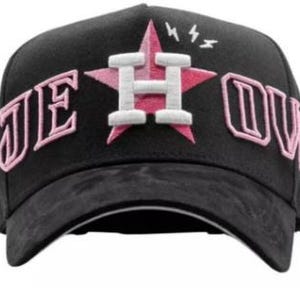 May include: Black baseball cap with a pink star and the letter "H" in white. The text "JE OUV" is embroidered in pink on the cap. The brim is black.