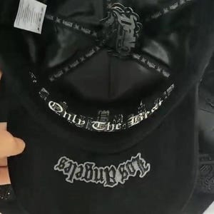 K&ouml;nnte beinhalten: Eine schwarze Baseballkappe mit einem inneren Schwei&szlig;band mit dem wei&szlig;en Text Only The Best und einem Kronenfutter mit einem schwarzen Logo-Aufn&auml;her. Die Unterseite des Schirms zeigt das Wort Los Angeles in grauer Stickerei im Gothic-Stil.