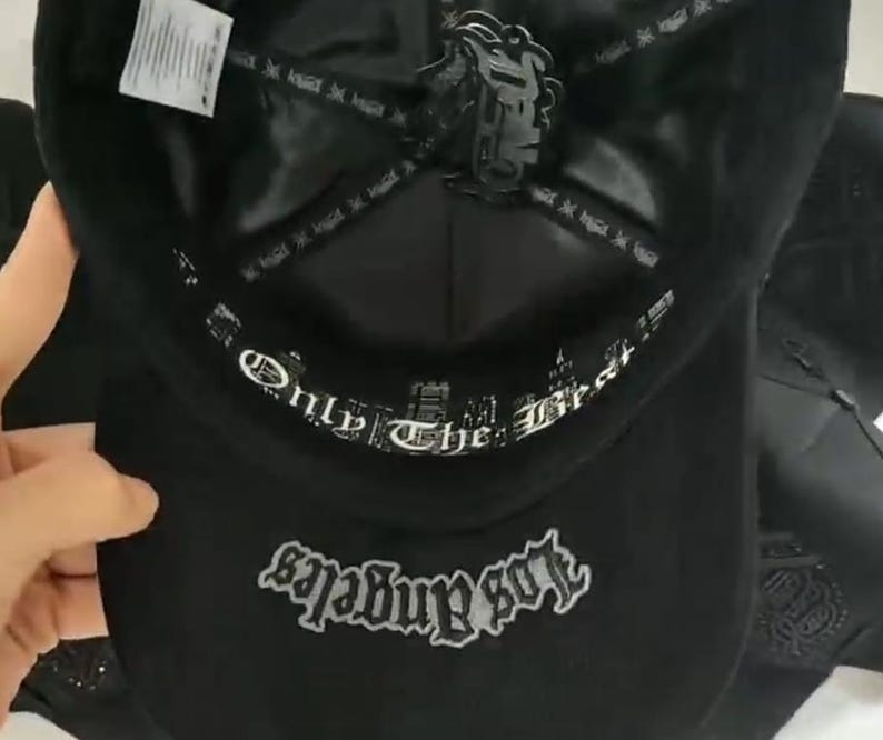 K&ouml;nnte beinhalten: Eine schwarze Baseballkappe mit einem inneren Schwei&szlig;band mit dem wei&szlig;en Text Only The Best und einem Kronenfutter mit einem schwarzen Logo-Aufn&auml;her. Die Unterseite des Schirms zeigt das Wort Los Angeles in grauer Stickerei im Gothic-Stil.