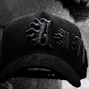 以下が含まれることがあります： 刺繍が施された黒い野球帽。帽子には、前面に装飾的な文字「H」のデザインと、その他の装飾要素があります。つばには追加の刺繍アクセントがあります。帽子は濃い無地です。