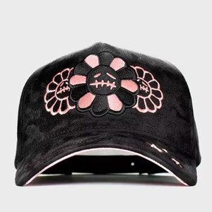 May include: A black velvet baseball cap featuring an embroidered design of three stylized flowers with alternating black and light pink petals. The central flower displays a stitched face with closed eyes and a straight line for a mouth.