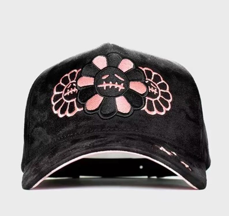 May include: A black velvet baseball cap featuring an embroidered design of three stylized flowers with alternating black and light pink petals. The central flower displays a stitched face with closed eyes and a straight line for a mouth.