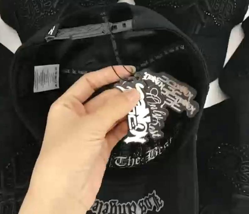 K&ouml;nnte beinhalten: Eine schwarze Baseballkappe mit verstellbarem Snapback-Verschluss, einem inneren Schwei&szlig;band mit dem Text The Best und einem wei&szlig;en Pflegeetikett. An der Kappe sind mehrere schwarz-wei&szlig;e Papieranh&auml;nger mit grafischen Textdesigns befestigt.