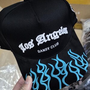 以下が含まれることがあります： 黒いベースボールキャップ。 白い刺繍で「Los Angeles」の文字。 つばには青い炎の刺繍があり、「DANDY CLUB」の文字も。 ファッションアクセサリーです。