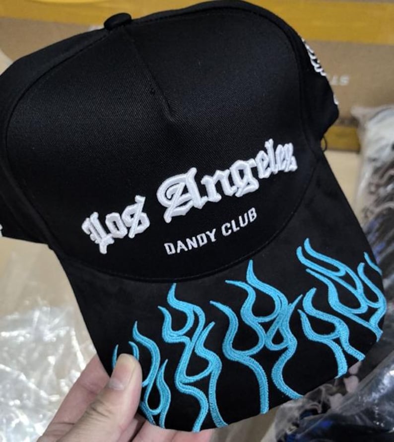 以下が含まれることがあります： 黒いベースボールキャップ。 白い刺繍で「Los Angeles」の文字。 つばには青い炎の刺繍があり、「DANDY CLUB」の文字も。 ファッションアクセサリーです。