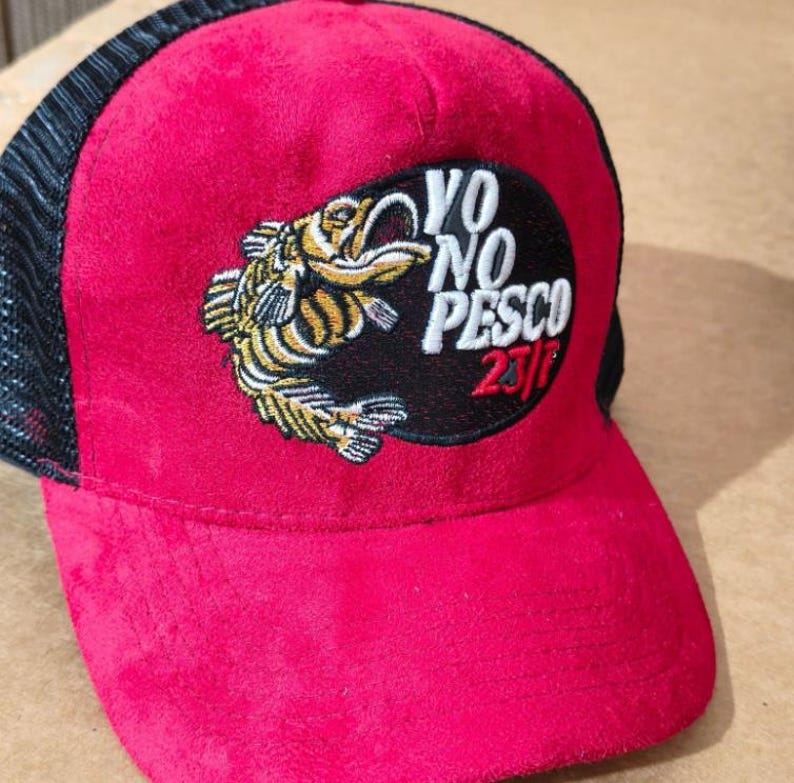 May include: A red baseball cap with a black mesh back. The front of the cap features a patch with a fish illustration and the text "VO NO PESCO 23/7". The cap is made of a soft, suede-like material.