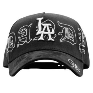 May include: Black baseball cap with white embroidered text that reads "LA Dad" in a gothic font. The cap has a curved brim and an adjustable strap at the back. The hat is made of a textured material.
