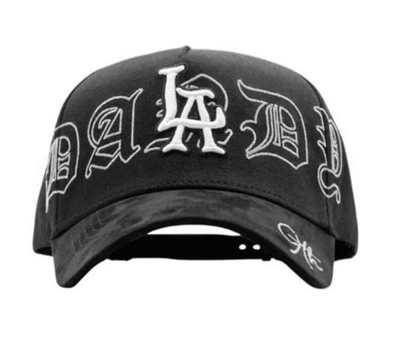 May include: Black baseball cap with white embroidered text that reads "LA Dad" in a gothic font. The cap has a curved brim and an adjustable strap at the back. The hat is made of a textured material.