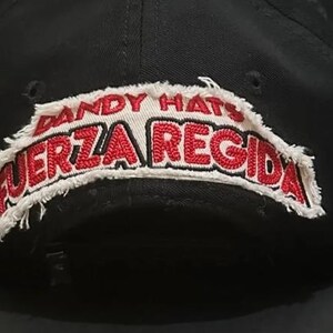 May include: A black baseball cap featuring a white fabric patch with frayed edges. The patch displays the text DANDY HATS in red and the text FUERZA REGIDA in bold red lettering. The cap has a structured crown and a small black adjustment strap.