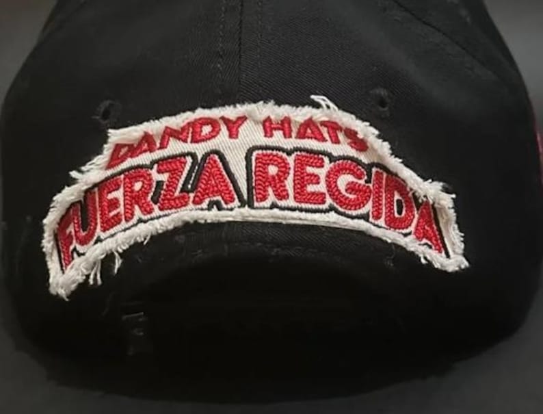 May include: A black baseball cap featuring a white fabric patch with frayed edges. The patch displays the text DANDY HATS in red and the text FUERZA REGIDA in bold red lettering. The cap has a structured crown and a small black adjustment strap.