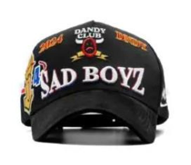 Może przedstawiać: Czarna czapka z daszkiem z białym napisem "SAD BOYZ" i "DANDY CLUB" nad małym emblematem. Czapka posiada r&oacute;wnież liczby "2024" oraz inne grafiki w kolorze złotym, czerwonym i niebieskim.
