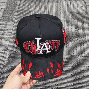 May include: A black baseball cap featuring the text "CHLA" in white and red embroidered lettering with rhinestone accents. The cap includes red cherry graphics on the sides and a red splatter pattern across the front brim and lower crown area.