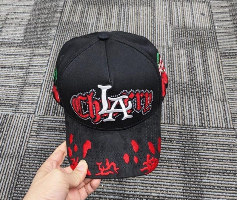 May include: A black baseball cap featuring the text "CHLA" in white and red embroidered lettering with rhinestone accents. The cap includes red cherry graphics on the sides and a red splatter pattern across the front brim and lower crown area.