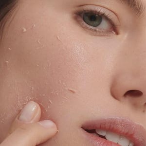 May include: A close-up view of a face showing skin with visible peeling or pilling texture. The skin appears light-toned with a natural finish, and a finger is touching the cheek area where small, thin flakes of product are lifting from the surface.