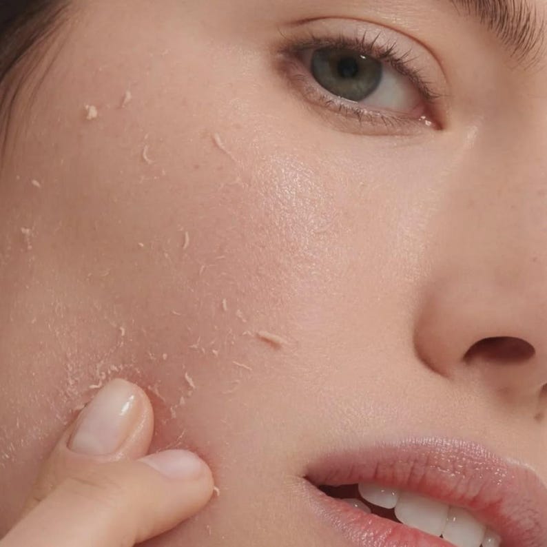 May include: A close-up view of a face showing skin with visible peeling or pilling texture. The skin appears light-toned with a natural finish, and a finger is touching the cheek area where small, thin flakes of product are lifting from the surface.