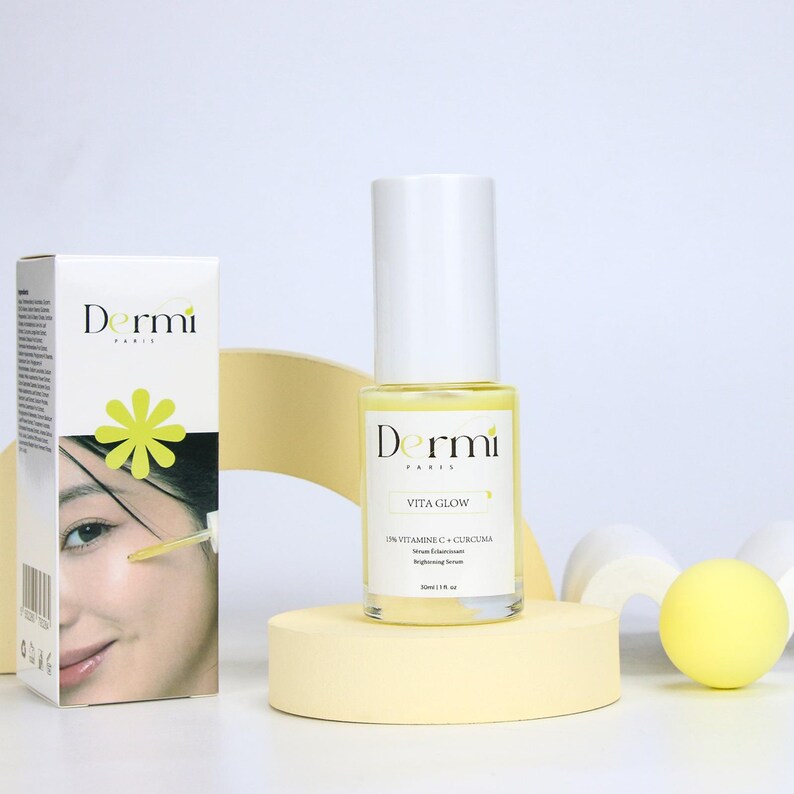 May include: A clear glass bottle of Dermi Paris Vita Glow brightening serum with a white cap, labeled 1 fl oz, standing next to its white rectangular box featuring a yellow flower icon and a photograph of a person applying the serum.