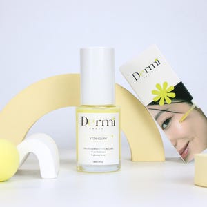 May include: A clear glass bottle of Dermi Paris Vita Glow 10% Vitamine C + Curcuma brightening serum with a white cap and label, containing 30ml | 1 fl oz. Next to it is a white product box featuring the brand name, a yellow flower, and a face.