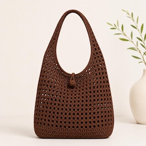 May include: A dark brown crochet hobo bag featuring an open square grid pattern throughout the body. The bag has a single integrated shoulder strap and a small, centered circular button closure at the top opening, crafted from matching brown yarn.