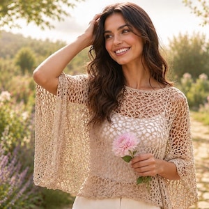 May include: A beige crochet poncho featuring an openwork pattern with wide, loose sleeves and a boat neckline. The garment is layered over a solid white top and is constructed from a lightweight, textured yarn with a delicate, net-like appearance.
