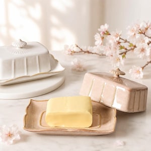 May include: A rectangular ceramic butter dish with a light pink glaze and a matching lid featuring a small knob handle. The dish has a fluted design along the sides and holds a block of yellow butter. A white ceramic butter dish is visible nearby.
