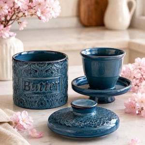 May include: A dark blue ceramic butter crock with an embossed floral pattern and the word Butter printed on the front. The set includes a cylindrical base, a matching lid with a knob handle, and an interior cup that sits on a small pedestal base.
