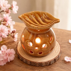 May include: A glossy, tan-colored ceramic oil burner with a leaf-shaped top dish and a rounded base featuring multiple circular cutouts. The burner is illuminated from within, casting a warm glow through the openings in its glazed surface.