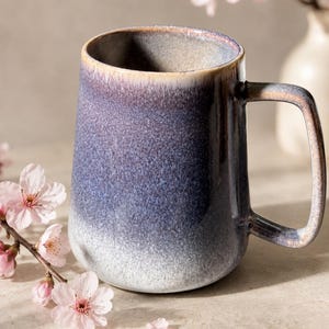 Könnte beinhalten: Ein Keramikbecher mit einer Farbverlaufglasur, die von einem tiefen Violett oben zu einem hellen Weiß an der Basis übergeht. Der Becher hat einen glatten, abgerundeten Körper und einen rechteckigen Henkel mit passender gesprenkelter Oberfläche.