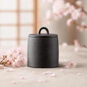 Può includere: Un contenitore cilindrico in ceramica nera con una finitura opaca e testurizzata, dotato di un coperchio abbinato con una piccola maniglia ad arco sulla parte superiore. Il recipiente ha un design semplice con pareti dritte e tinta unita.