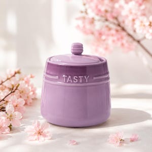 May include: A lavender ceramic storage jar with a matching lid, featuring the word TASTY embossed on the front. The container has a smooth, glossy finish and a slightly tapered cylindrical shape with horizontal ridged detailing near the top rim.