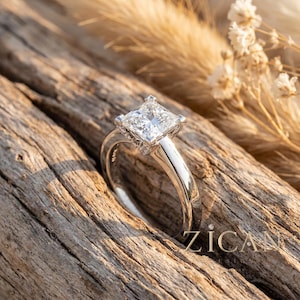 May include: A silver-toned ring featuring a square princess-cut centre stone held by four prongs. The band is smooth and polished, with small accent stones set along the gallery. The text ZICAN is visible in the lower right corner of the image.