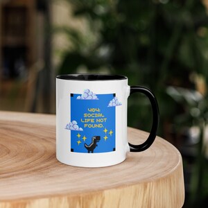 Può includere: Tazza in ceramica bianca con manico e bordo neri. La tazza presenta un quadrato blu con nuvole pixelate, stelle e una grafica di dinosauro. Il testo sulla tazza recita "404: SOCIAL LIFE NOT FOUND."