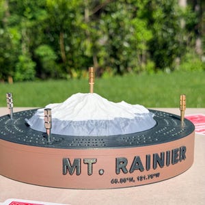 May include: A circular cribbage board featuring a 3D-printed white and gray model of Mt. Rainier in the center. The base is tan with the text "MT. RAINIER" and "46.85°N, 121.76°W" printed on the side. Four metal pegs are inserted into the board.