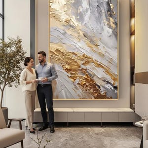 Op de afbeelding: Groot abstract schilderij met gouden, grijze en witte getextureerde penseelstreken. Het kunstwerk is omlijst in een gouden rand en hangt boven een lage kast. Een stel staat voor het schilderij in een moderne woonkamer.