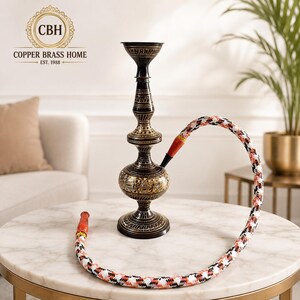 May include: A black metal hookah with intricate gold-toned etched patterns on the base and stem. The hookah features a long, flexible hose with a white, black, and orange woven diamond pattern and solid orange end pieces for a distinct appearance.
