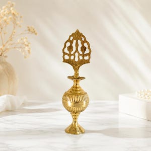 May include: A gold-colored metal perfume bottle featuring a rounded, ribbed body and a tall, ornate, openwork stopper with a pointed top. The bottle stands on a flared pedestal base and has a polished, reflective finish throughout its surface.