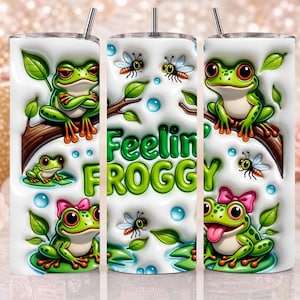 May include: A white cylindrical tumbler featuring a repeating pattern of green frogs, bees, and leaves. The text "Feelin FROGGY" is printed in large green letters. The design includes blue water droplets and a frog wearing a pink bow.