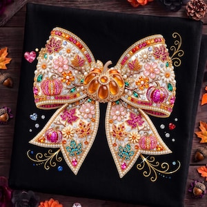 May include: A black t-shirt featuring a large, ornate bow graphic decorated with colorful rhinestones, pearls, and gems. The design includes autumn-themed motifs such as pumpkins, maple leaves, and flowers in shades of orange, pink, and teal.