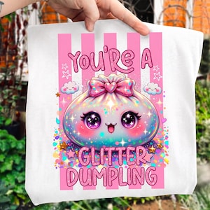 May include: A white tote bag featuring a graphic of a cute, iridescent dumpling with large purple eyes, a pink bow, and sparkles. The text reads "YOU'RE A GLITTER DUMPLING" in pink letters against a background of vertical pink stripes, small clouds, and stars.