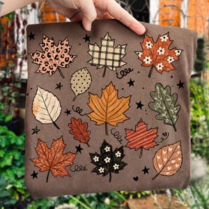 Op de afbeelding: Een bruin t-shirt met een grafische print van diverse herfstbladeren in verschillende patronen, waaronder luipaardprint, ruitjes, strepen en bloemmotieven, afgewisseld met kleine zwarte sterretjes en krullende lijnen.