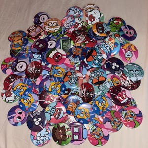 Puede incluir: Una colección de botones redondos y coloridos con varios diseños de personajes de dibujos animados. Los botones presentan una variedad de colores, incluyendo azul, rosa, rojo y verde. Los diseños incluyen caras, formas abstractas y otras ilustraciones caprichosas.