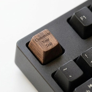 May include: A single brown wooden keycap with a visible wood grain texture, featuring the engraved text "Customize Your Text" in a dark font, installed on a black mechanical keyboard among standard black plastic keys.