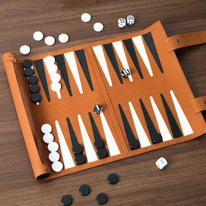 Pode incluir: Um conjunto de backgammon em couro castanho com peças brancas e pretas e dados. O tabuleiro está aberto, exibindo a superfície de jogo com triângulos brancos e pretos alternados. Vários dados estão espalhados no tabuleiro e algumas peças estão por perto.