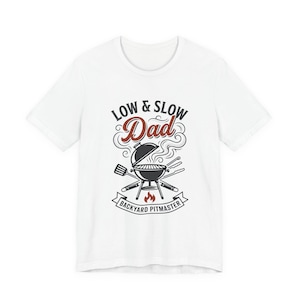 Pode incluir: T-shirt branco com um gráfico temático de churrasco. O design apresenta uma grelha, utensílios de cozinha e o texto "LOW & SLOW Dad BACKYARD PITMASTER" em estilo vintage. Um presente perfeito para os amantes de churrasco.